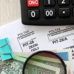 Kalkulator, formularze PIT, pieniądze i lupa na drewnianym tle.